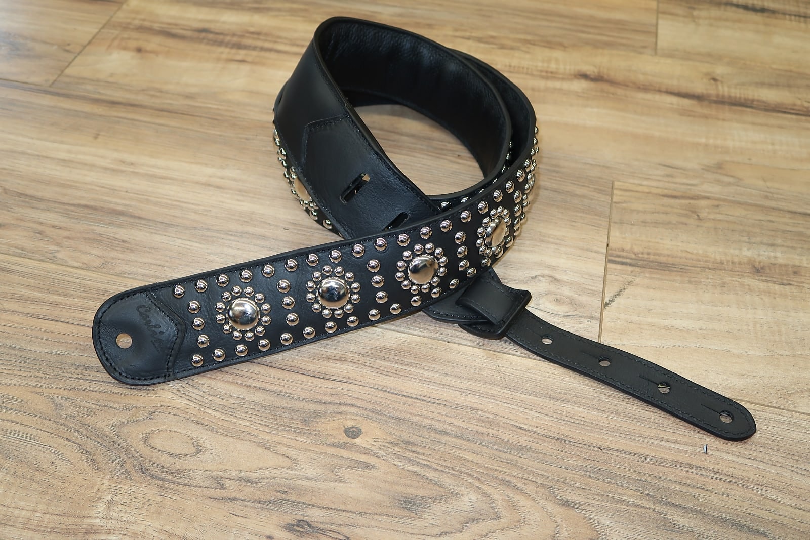 Black leather guitar strap with silver studded detailing on a wood floor